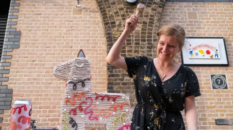 A woman standing next to a piñata in the shape of a horse, made of gingerbread. She is holding a small hammer above it and is smiling
She is wearing a black dress with small flowers on it. the horse is covered in iced beading, with black, orange, green and brown iced squiggles along its side.