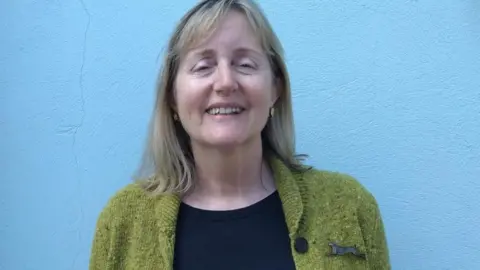 MANX DEAF SOCIETY Heather Hurt, a woman with a friendly smile, she has a fringe and brown/grey hair, stood in front of a blue wall.