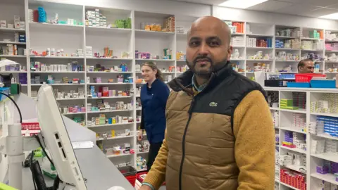 A busy pharmacy showing medicine stacked on the shelves. a man in a yellow jumper and yellow gilet stands behind the counter
