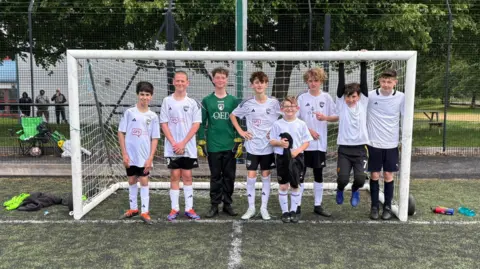 Simon Sibley Eight teenage boys stand under a goalpost, wearing white football shirts apart from one who wears a green shirt and goalkeeper gloves.