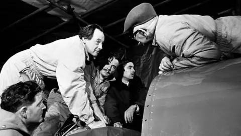 Getty Images Black-and-white photograph of Donald Campbell talking to engineers as the gather around Bluebird. He has dark hair, brushed back from his face. He is wearing overalls.