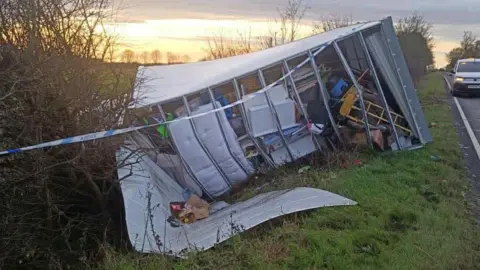 A large white lorry full of belongings tipped on its side on a roadside grass verge. 