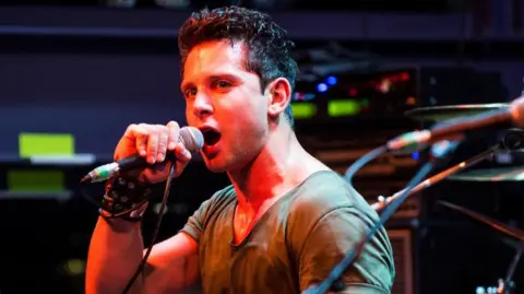 Man with brown hair sings into microphone on stage with red lighting shining down on him. He wears a tan t-shirt and black punk wristband.