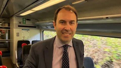 Matt Barnes is aboard a train smiling at the camera. He has short dark hair and is wearing a dark grey jacket over a purple and white patterned shirt with a navy and white printed tie.
