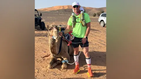 Marc Lockwood Marc Lockwood pictured with a camel. The camel is sat on its legs, while Marc is holding a leash. Marc is wearing a white hat, sunglasses and rucksack. He is also wearing a green t-shirt, black shorts and orange trainers.