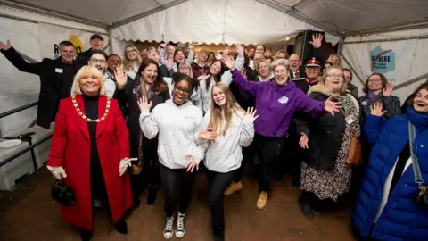 A big group of people smiling and waving at the camera in the inside of a tent.