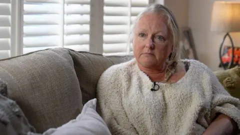 A woman with blonde hair and wearing a white jumper sits on a grey sofa in the living room of a house.