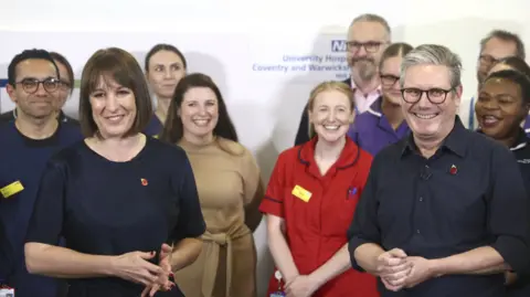 PA Media Chancellor of the Exchequer Rachel Reeves on a hospital visit with Prime Minister Sir Keir Starmer. Both are wearing navy clothes and small poppy badges, and smiling, and there is a team of medical staff behind them