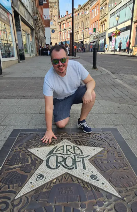 Suppplied A man kneeling down to touch an engraved star with the words LARA CROFT on the floor