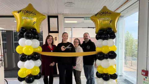 Three women and one man stand in a row behind a yellow ribbon and balloon tower with gold stars on top. The woman in the middle is dressed all in black and holding a large scissors