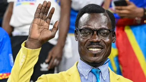 Michel Nkuka Mboladinga dressed as Patrice Lumumba in a yellow suit jacket and blue cheque shirt and tie lifts his hand as if waving in the stands at Afcon.