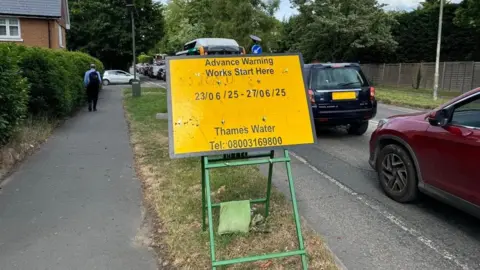 A yellow sign gives motorists advance warning of Thames Water works in Merrow in Guildford. Cars queue up alongside. In the background a man walks along a pavement.
