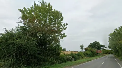 Google A street view image of a field taken from a two-lane road. A brown street sign can be seen in the distance. Trees and bushes line the road.