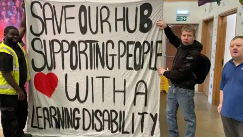 People holding a white banner with the words "Save our hub, supporting people with a learning disability"