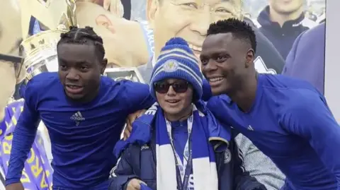 Leicester City Two football players wearing blue undershirts with their arms around a boy, wearing a woolly hat and sunglasses
