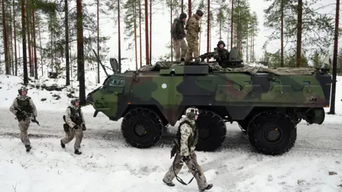 An armoured vehicle on a snow-covered road. There are three soldiers on top of the tank and three others walking along side it. 