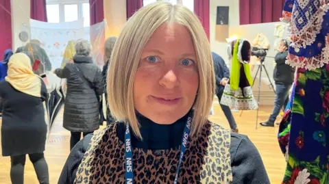 BBC A woman smiles at the camera, she stands in Fearon Hall Community Centre with Afghan women and artwork behind her 