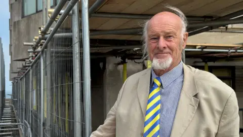 Hannah Roe/BBC Simon Peter Pengelly, wearing a light-coloured jacket, striped shirt and blue-and-yellow striped tie, stands beside metal fencing and scaffolding surrounding repair works at a seafront building. The exterior of the building and temporary support structures can be seen behind him.