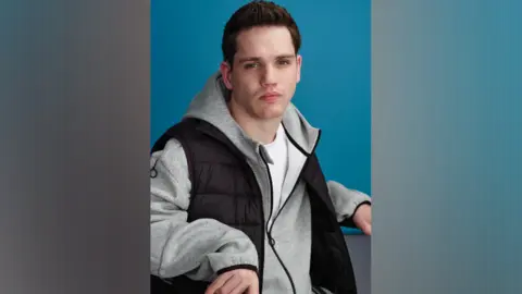 Primark Elliott Caswell looks directly at the camera, wearing a grey hooded top and a black bodywarmer and white t-shirt. He has a serious expression and is pouting slightly. 