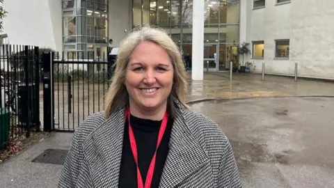 Hannah Knowles is wearing a black top and grey coat and has a red lanyard around her neck. She is standing at the school gates and there are school buildings behind her.