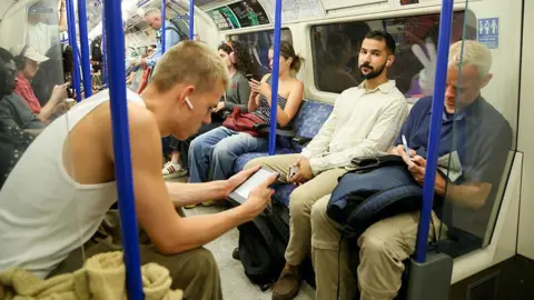 An underground carriage with people entertaining themselves without phones. One man is reading a Kindle and another is doing a crossword with a pen. 