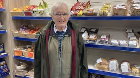 A man is stood in the middle of the shot - he's wearing glasses and looking at the camera. He also has on a green coat and blue jumper. Around him is fruit and vegetables, as well as baked goods all on shelves.