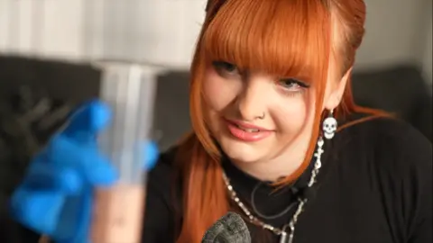 A woman with orange hair holds a clear tube with her right hand. She is wearing a blue glove