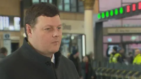 Mark has short brown hair. He is wearing a grey jacket.