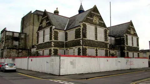 A view of an old Victorian building, with metal sheets where the windows should be and hoarding around the edge of the building perimeter.