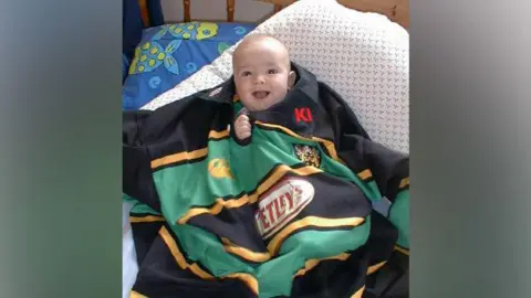 Supplied Matthew Sansome as an infant. He is on a bed and has a black, green and gold Northampton Saints jersey draped over him, with his head and one hand peeping out of the collar.