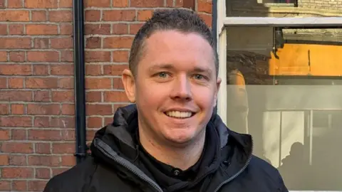 Close shot of dale lee, he has short brown hair and is smiling at the camera.He is pictured against a red brick wall and is wearing a black coat