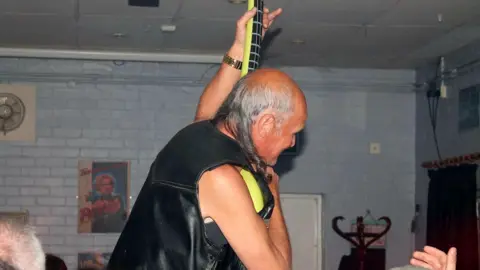 Cottenham Rock Club Brian Dean with short white hair, wearing a leather jacket and "playing" a green inflatable guitar, which he is holding vertically. He is in a pub or similar and the hand of an audience member is raised to the right of the picture. There is a hatstand in the background and pictures on a brick wall that has been painted light blue.