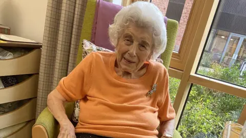 An elderly lady sat in a green chair wearing an orange jumper