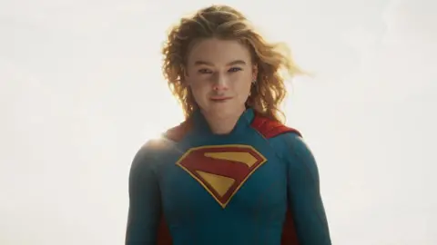 DC Studios Milly Alcock, wearing the blue Supergirl suit complete with red cape and red S emblazoned on the chest, stands against a hazy, sunny sky and stares down the camera with a wry smile on her face.