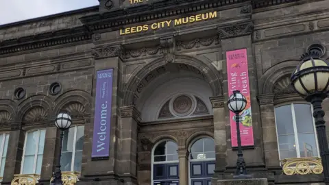 The entrance to Leeds City Museum - an old stone building with its name emblazoned in gold above the doors, which are glassed and blue.