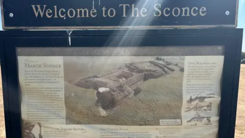 SUPPLIED An impression of a fort from the 1600s, a cannon is being fired with much smoke and some soldiers can be seen, it is on grass land with a moat cut in surrounding the fort. The is text explaining the sconce surrounding the picture. 