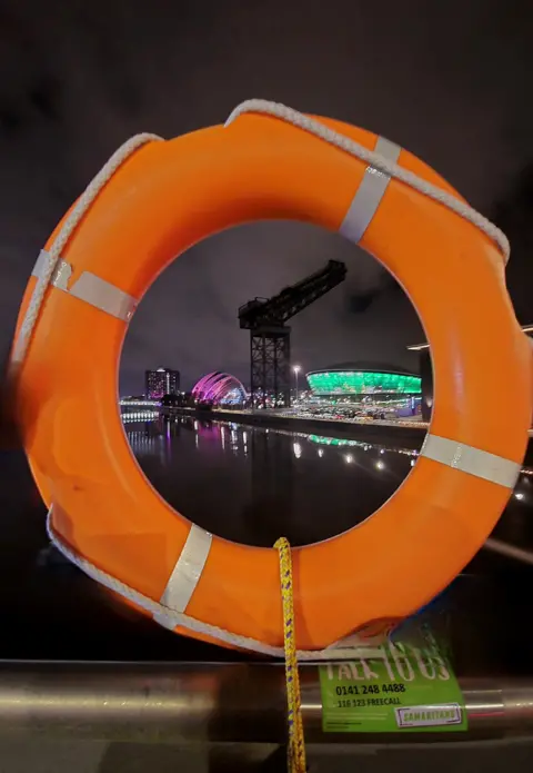 Jill Ferguson A circular orange life ring on the riverside, with a large crane and large arena visible through the space in the centre of the aid