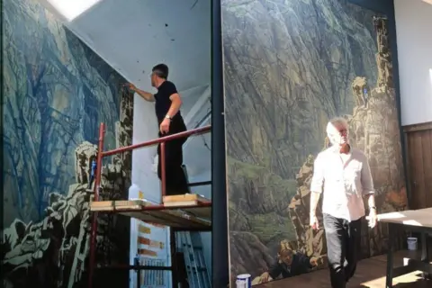 Heaton Cooper Studio A spilt of two pictures. On the left is Julian Cooper painting Scafell Crag in 2001, and on the right is Julian Cooper with the painting 20 years later in 2021