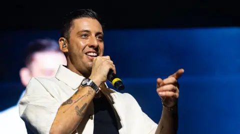 Musician Example wearing a black to and cream shirt, holding a microphone in his right hand and pointing with his left hand whilst performing