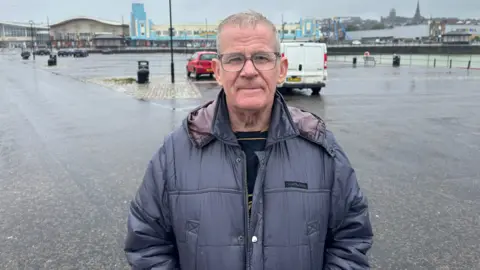 Sean Martin wearing glasses and a navy waterproof coat, looking at the camera, with his hands in his pockets, standing in a car park. It looks wet and it is raining. 