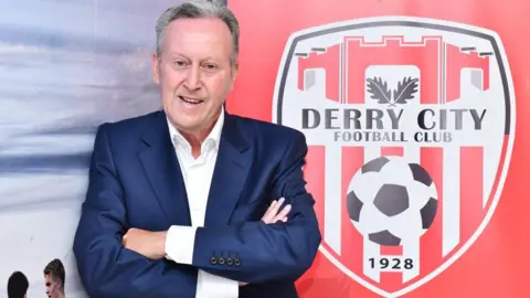 A man in a navy suit jacket and white shirt stands with his arms folded. 
He is beside a large red and black sign featuring the Derry City Football Club crest. 