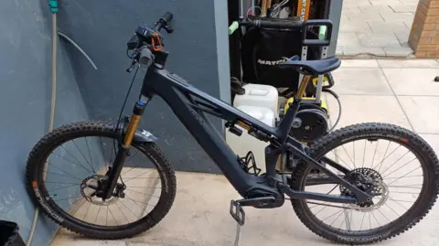 PA Media A black e-bike in a yard with beige Indian stone tiles confiscated by police officers.