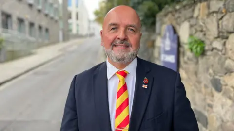 Man in his late 50s wearing a blue suit and a yellow and red striped tie. he is bald and has a white and grey beard 