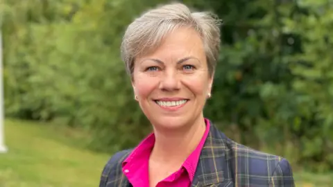 A smiling grey-haired woman standing on a lawn and in front of bushes, wearing a tartaned blazer and pink shirt.