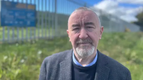 Brian Higginson has grey hair and a grey beard. He is standing outside during the day in front of grass in front of a metal fence. He is wearing a shirt under a jumper and blazer.