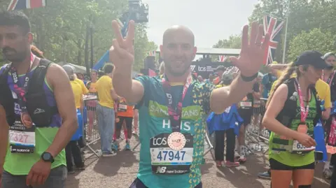 Chris at the finish line of the London Marathon, wearing his medal. His hands are aloft, one in a peace sign. Other finishers are around him.