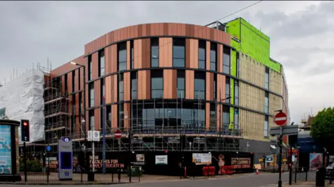 City of Wolverhampton Council The new building curves round the corner of two streets in the city. It is orange, green and glass with some scaffolding still on the exterior and hoardings on the street level storey of the building.
