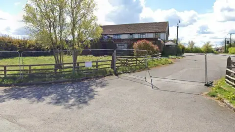 A two-storey building cordoned off by a metal fence. There are trees and greenery surrounding the site and the sky is blue. 