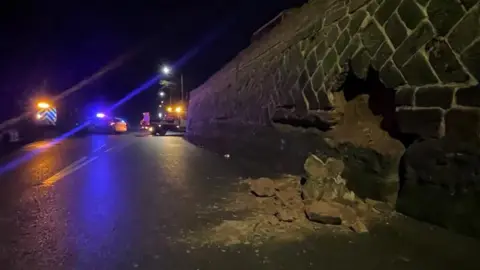 A section of collapsed wall strewn onto the road with emergency services blocking traffic in the distance