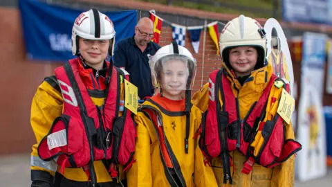 Three children wearing lifeboat crew gear, including white helmets, red lifejackets and yellow oilskins, smile as they stand on a promenade. Behind them, colourful flags and banners are attached to a red brick wall, while a man wearing a dark polo shirt and glasses looks down at a table.